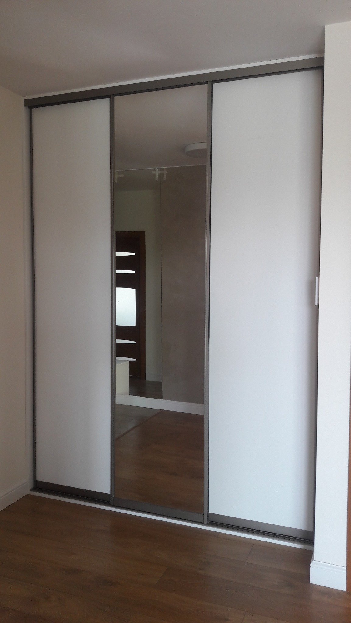 Wnętrze z szafą przesuwną, której środkowe drzwi są lustrem odbijającym pokój z drewnianą podłogą i drzwiami. Minimalistyczny design, szara rama.