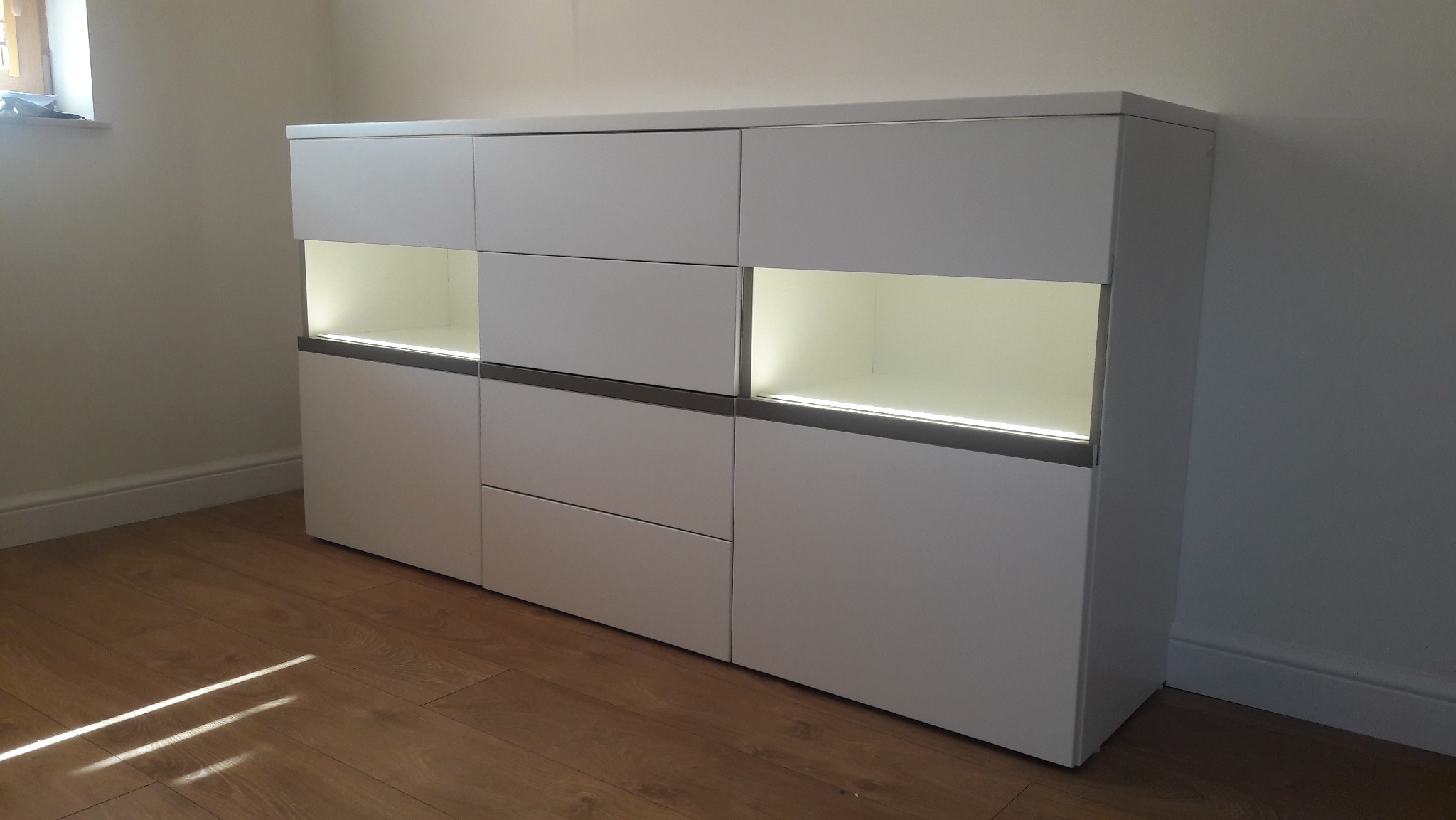 Biała komoda z szufladami i szafkami, podświetlana od wewnątrz, stoi na drewnianej podłodze w jasnym pomieszczeniu. Minimalistyczny design.