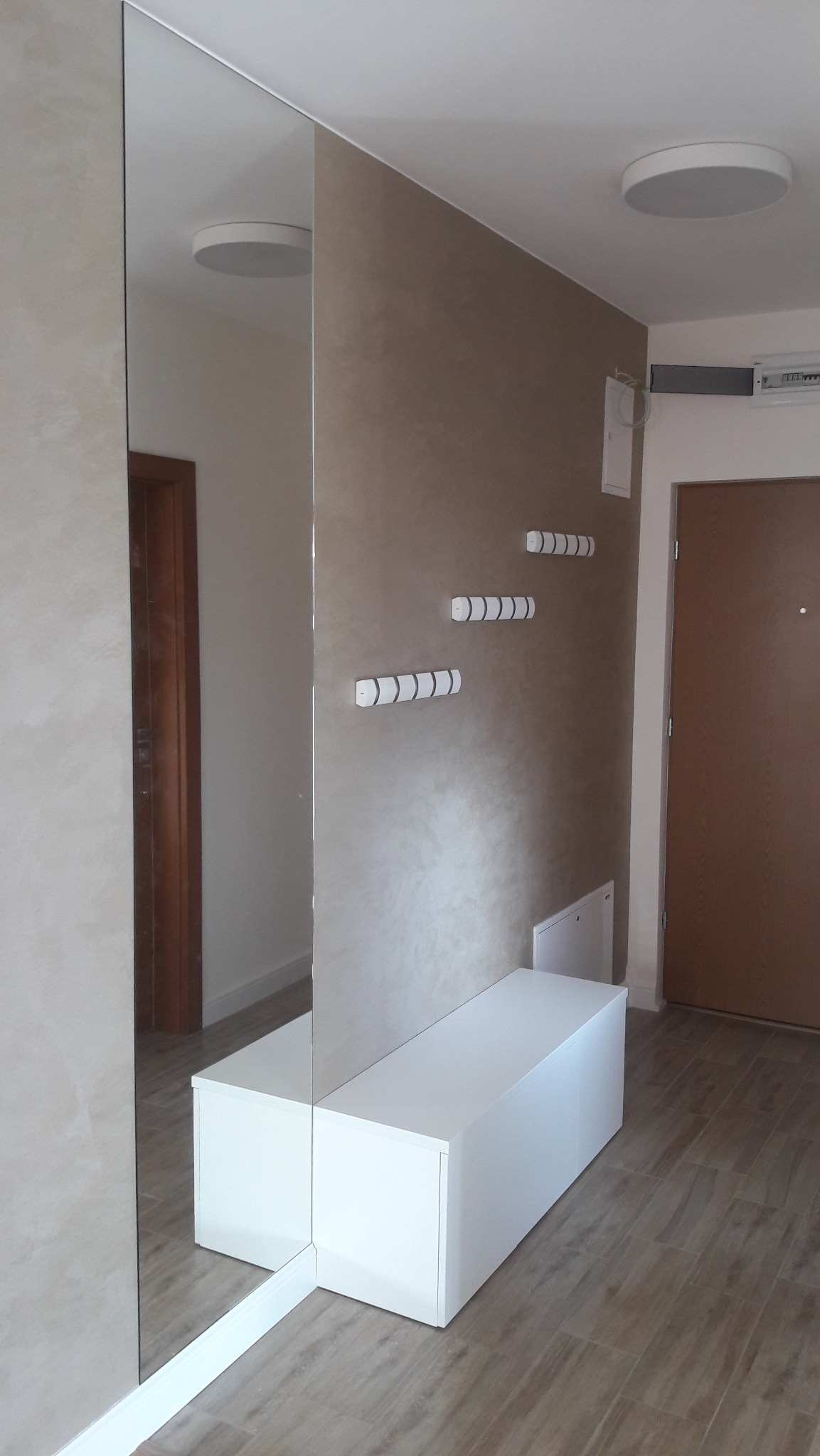 Nowoczesna szafa wnękowa z lustrem, białe fronty, minimalistyczne wieszaki na ścianie. Całość utrzymana w jasnej kolorystyce, z drewnianą podłogą.