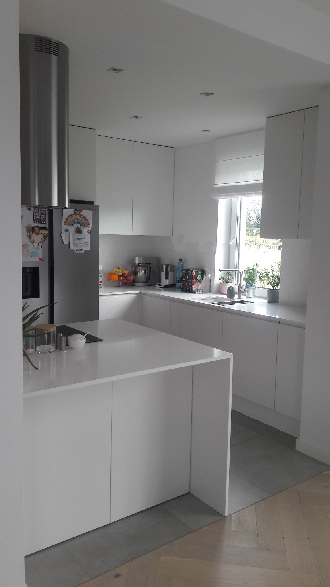 Jasna kuchnia z wyspą i okapem. Białe fronty szafek, szare płytki podłogowe i drewniana podłoga w jodełkę. Na blacie widoczne akcesoria kuchenne i dekoracje.