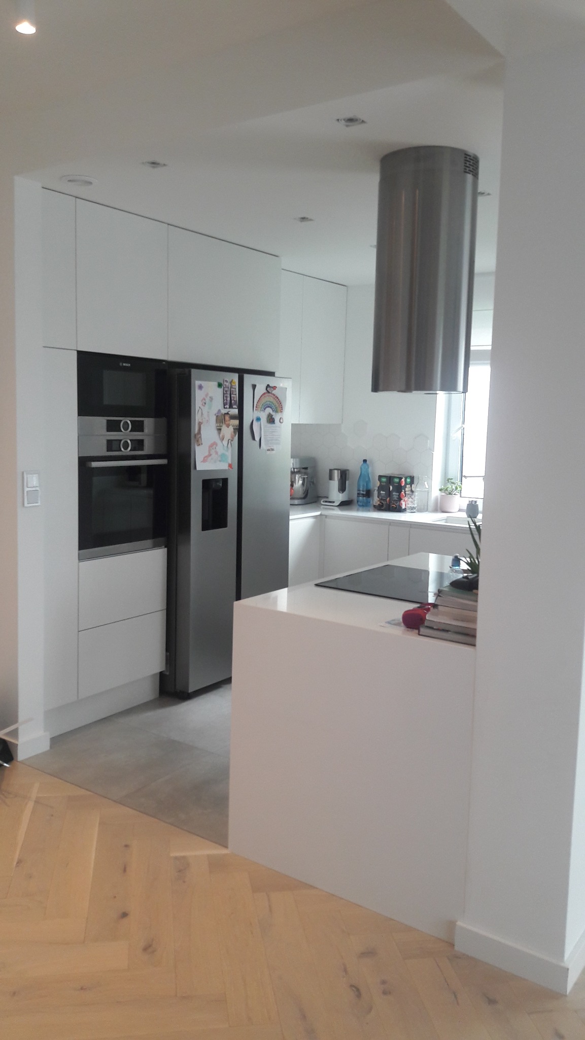 Nowoczesna, biała kuchnia z wyspą i sprzętem AGD: lodówka side-by-side, piekarnik w zabudowie. Parkiet w jodełkę. Minimalistyczny design, stonowana kolorystyka.