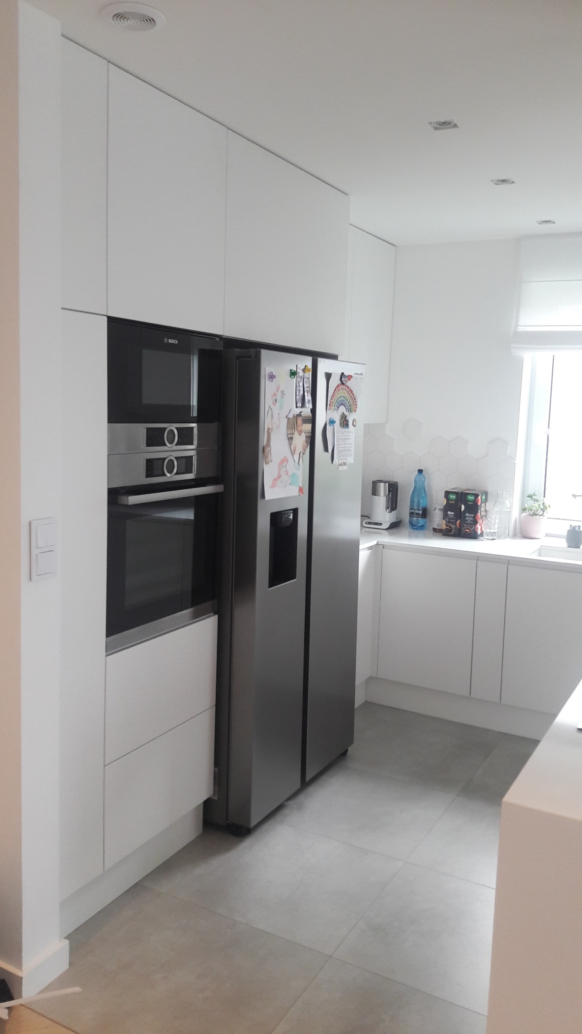 Nowoczesna kuchnia: białe szafki, zabudowana lodówka side-by-side, piekarnik Bosch, szare płytki podłogowe. Minimalistyczny design z akcentami w postaci magnesów na lodówce.