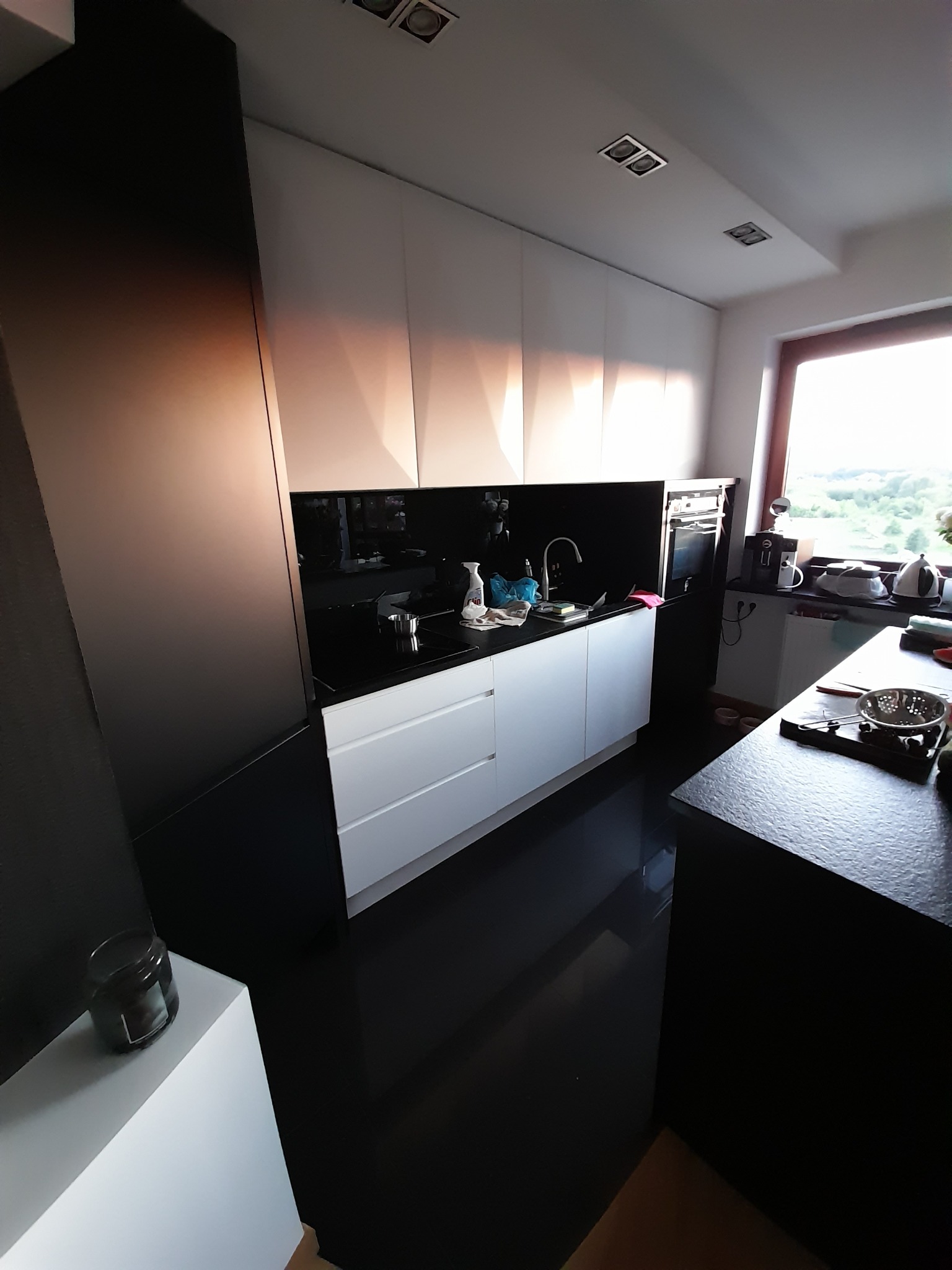 Nowoczesna kuchnia z białymi szafkami i czarnym blatem, oświetlona światłem dziennym wpadającym przez okno. Minimalistyczny design i czyste linie.