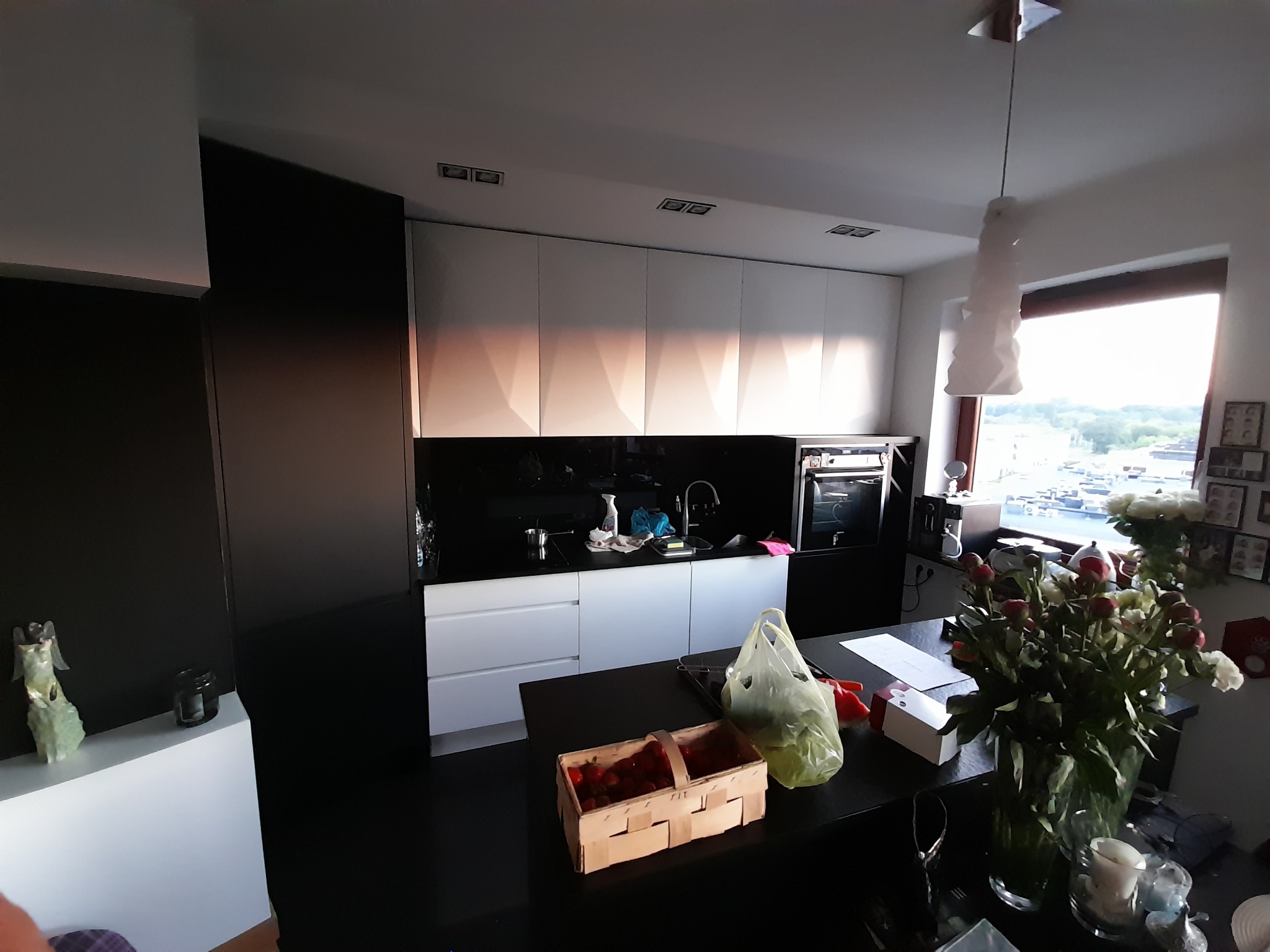 Nowoczesna kuchnia z białymi szafkami i czarnym blatem, oświetlona naturalnym światłem z okna. Na blacie koszyk truskawek i bukiet kwiatów.