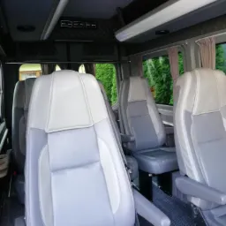 Wnętrze busa z szarymi, sk&oacute;rzanymi fotelami pasażerskimi, widok na okna z zasłonkami i panele sterowania oświetleniem.
