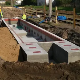 Budowa betonowego kanału technicznego przy torach kolejowych, widoczni pracownicy w odblaskowych kamizelkach i kaskach, wykop ziemny, niebieskie rury w tle.