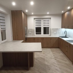 Ewemi Meble - Grzegorz Zygmunt - Nowoczesna kuchnia z drewnianymi frontami, jasnym blatem i oświetleniem LED. Widoczny stół/wyspa kuchenna i okno z żaluzjami. Ujęcie z perspektywy szerokokątnej.