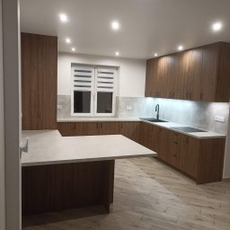 Ewemi Meble - Grzegorz Zygmunt - Nowoczesna kuchnia z drewnianymi szafkami, jasnym blatem i wyspą. Uwagę przyciąga oświetlenie LED pod szafkami i drewnopodobna podłoga.