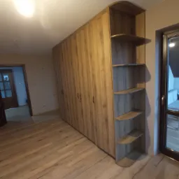 Szafa na wymiar z drewnianymi frontami i otwartymi półkami na boku, dopasowana do wnętrza. Widok na salon przez otwarte drzwi.