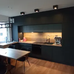 Nowoczesna kuchnia z czarnymi frontami i drewnianym blatem, oświetlona punktowymi lampami sufitowymi. Widoczny stół z krzesłami i dekoracyjna lampa wisząca.