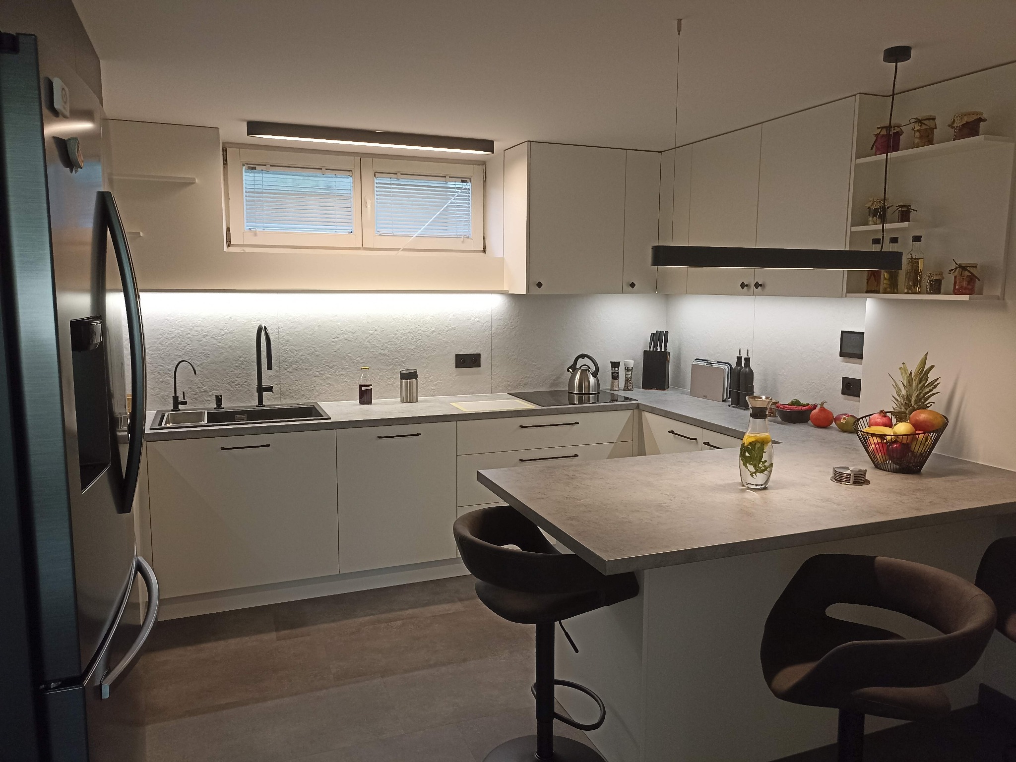 Nowoczesna kuchnia w bieli z szarym blatem, oświetleniem LED podszafkowym i wyspą z hokerami. Na blacie owoce i napój, minimalistyczny design.