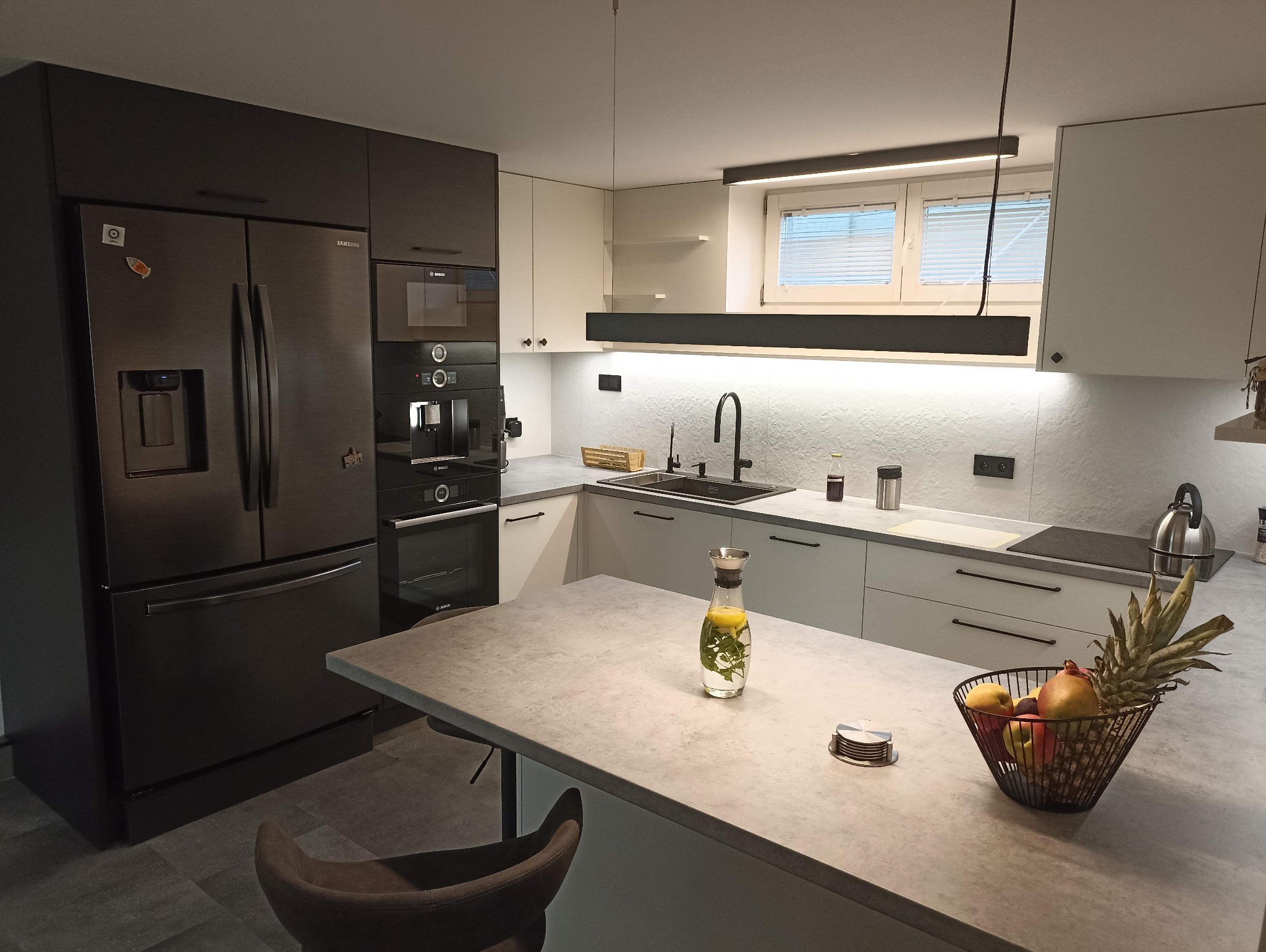 Nowoczesna kuchnia z czarnymi frontami i sprzętem AGD Bosch, oświetleniem LED, blatem w kolorze betonu i owocami na wyspie. Minimalistyczny design.