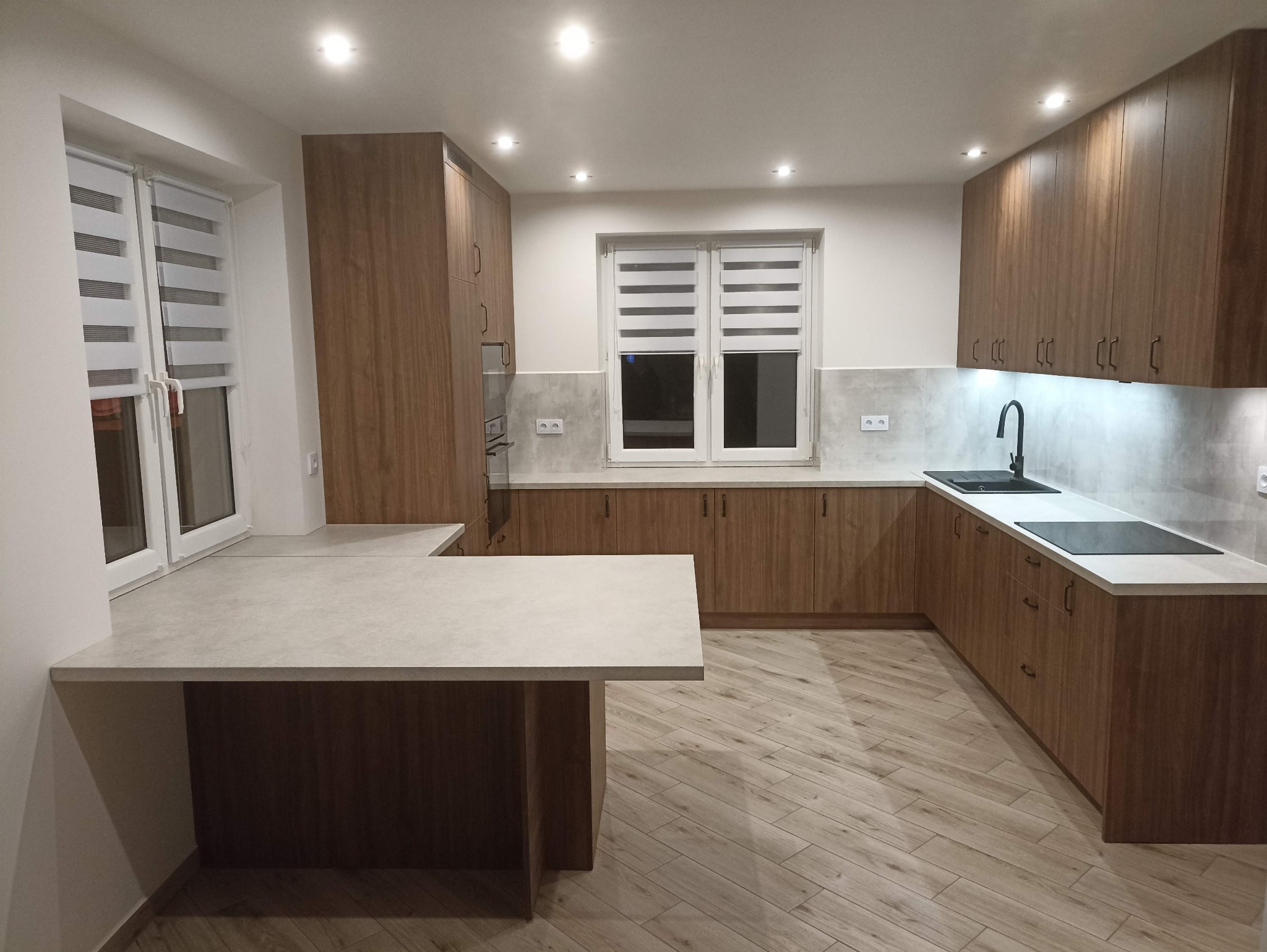 Nowoczesna kuchnia z drewnianymi frontami, jasnym blatem i oświetleniem LED. Widoczny stół/wyspa kuchenna i okno z żaluzjami. Ujęcie z perspektywy szerokokątnej.