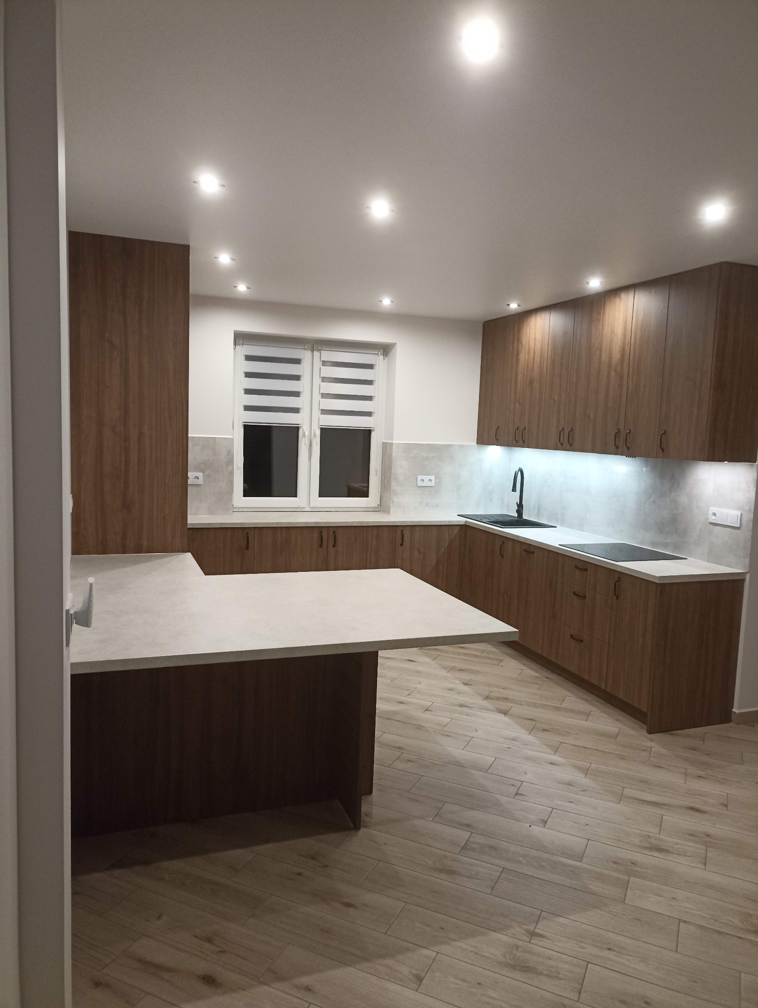 Nowoczesna kuchnia z drewnianymi szafkami, jasnym blatem i wyspą. Uwagę przyciąga oświetlenie LED pod szafkami i drewnopodobna podłoga.