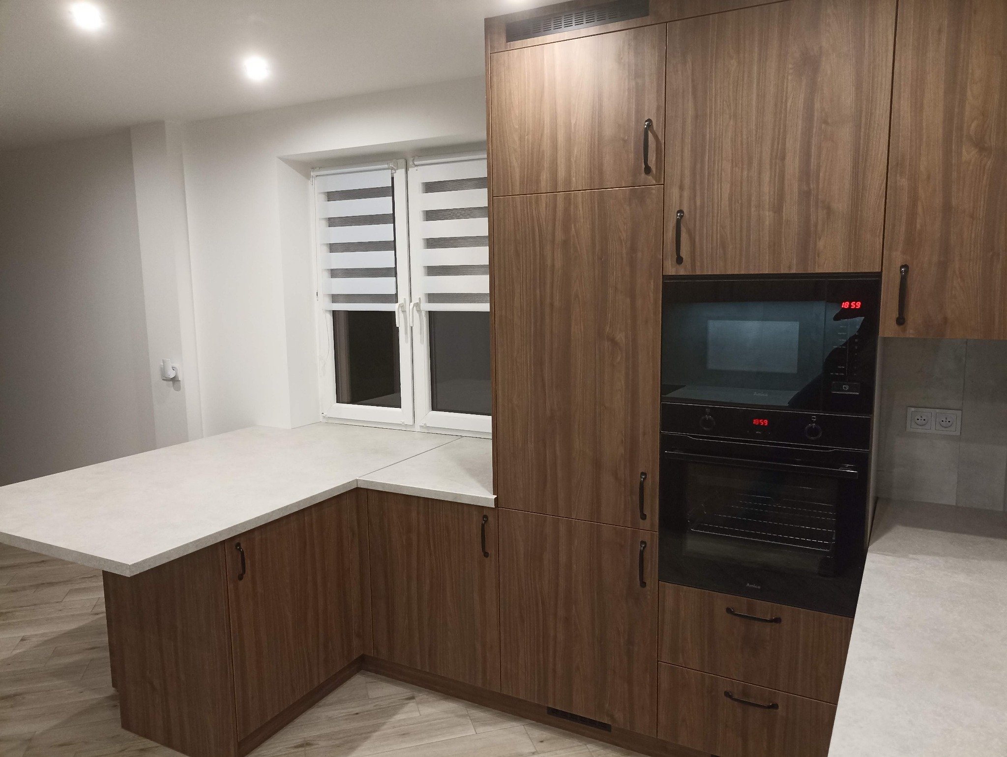 Nowoczesna kuchnia z drewnianymi frontami, jasnym blatem i zabudowanym piekarnikiem. Widoczne okno z roletami. Minimalistyczny design.