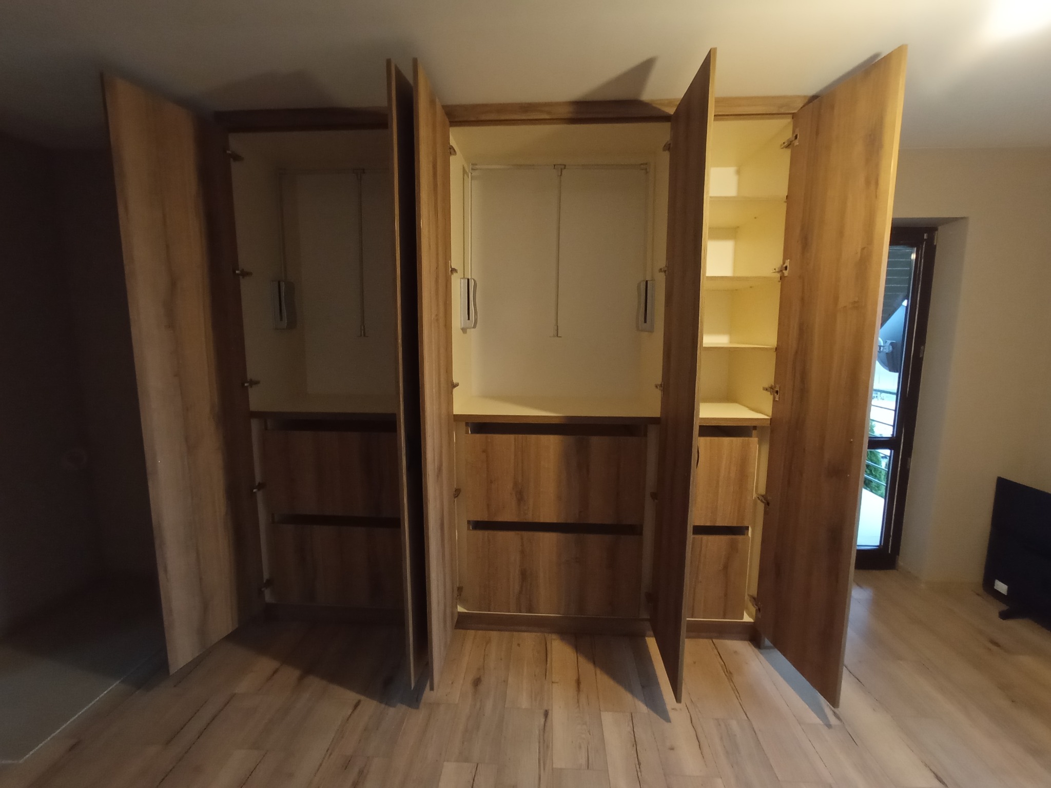 Drewniana szafa wnękowa z otwartymi drzwiami, ukazująca półki i drążki na ubrania. Widoczne szuflady i drewniana podłoga w pokoju.