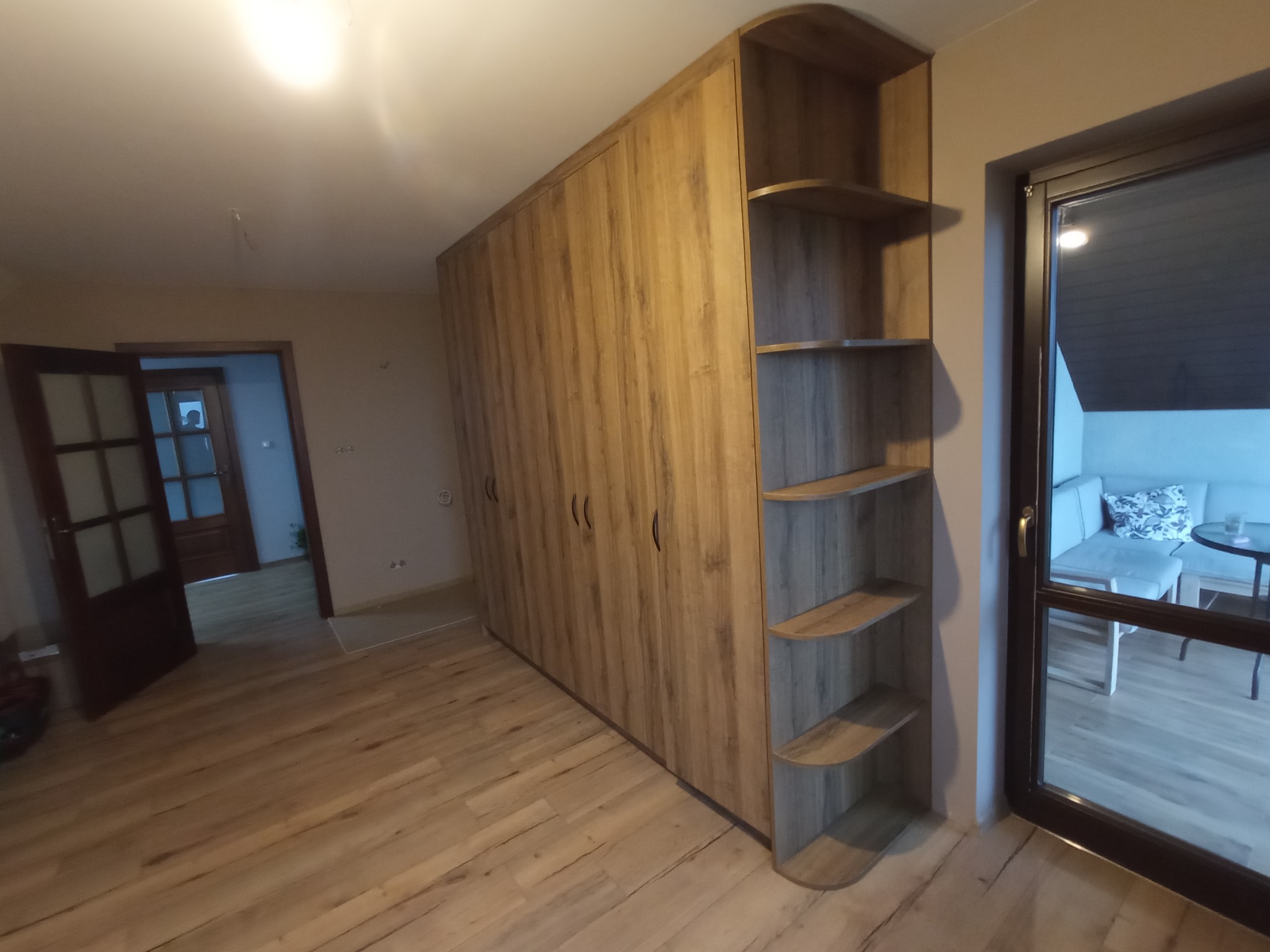 Szafa na wymiar z drewnianymi frontami i otwartymi półkami na boku, dopasowana do wnętrza. Widok na salon przez otwarte drzwi.