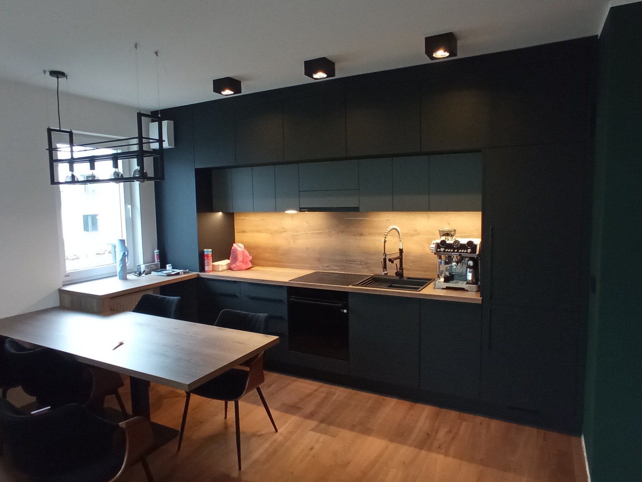 Nowoczesna kuchnia z czarnymi frontami i drewnianym blatem, oświetlona punktowymi lampami sufitowymi. Widoczny stół z krzesłami i dekoracyjna lampa wisząca.