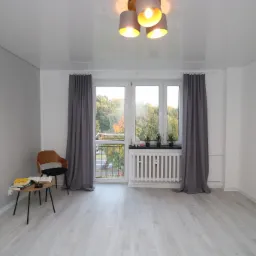 Jasne, puste pomieszczenie po remoncie z drewnianą podłogą, białymi ścianami, oknem z szarymi zasłonami, grzejnikiem, stolikiem z książkami i designerską lampą sufitową.