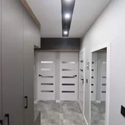 Nowoczesny przedpokój w szarościach z szafą w zabudowie, lustrem i białymi drzwiami z poziomymi przeszkleniami, oświetlenie sufitowe w czarnej wnęce.