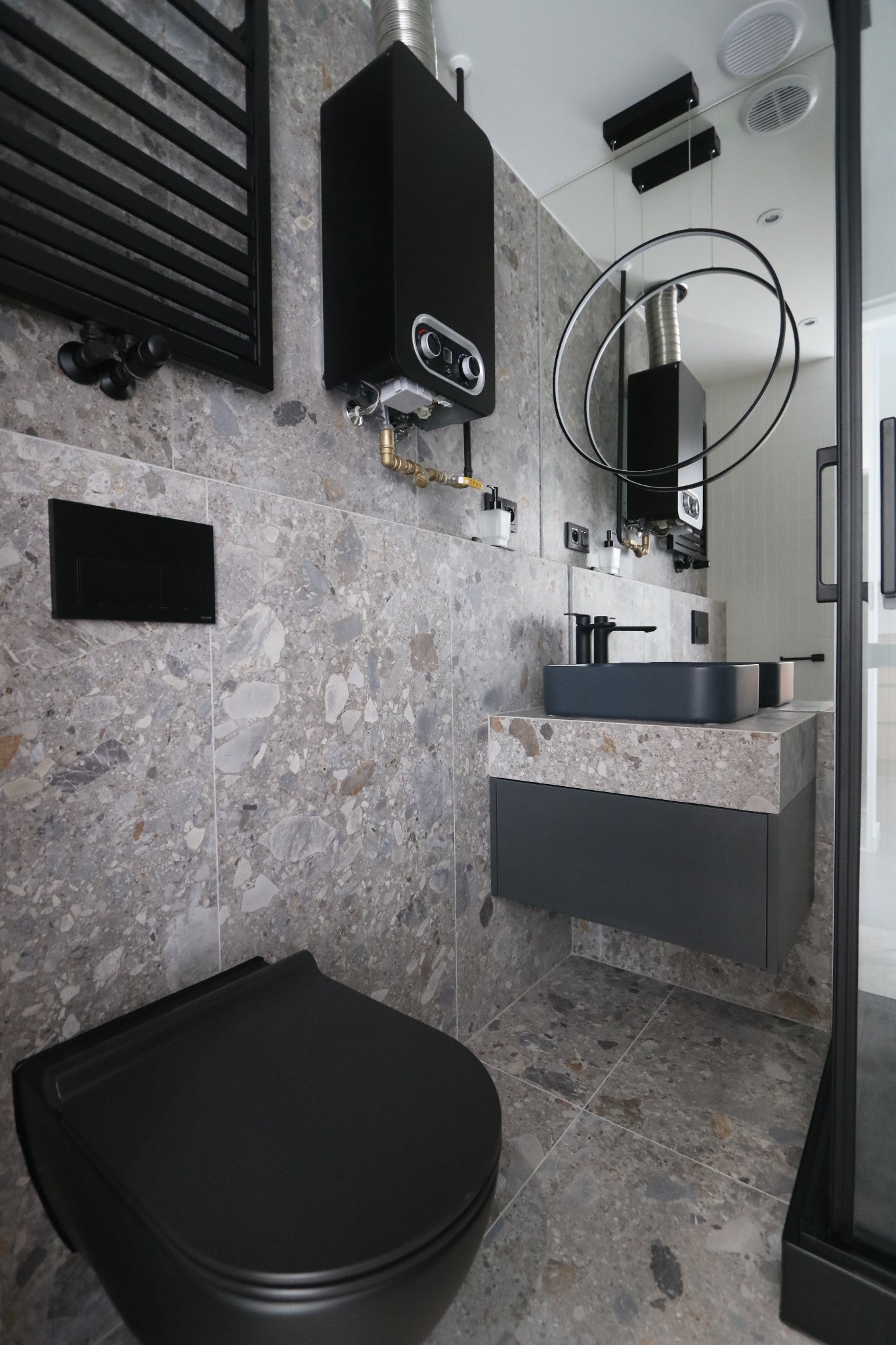Nowoczesna łazienka w Jastrzębiu-Zdroju z szarymi płytkami imitującymi kamień, czarną armaturą, podwieszaną toaletą i grzejnikiem drabinkowym, oraz podwieszaną szafką z umywalką.