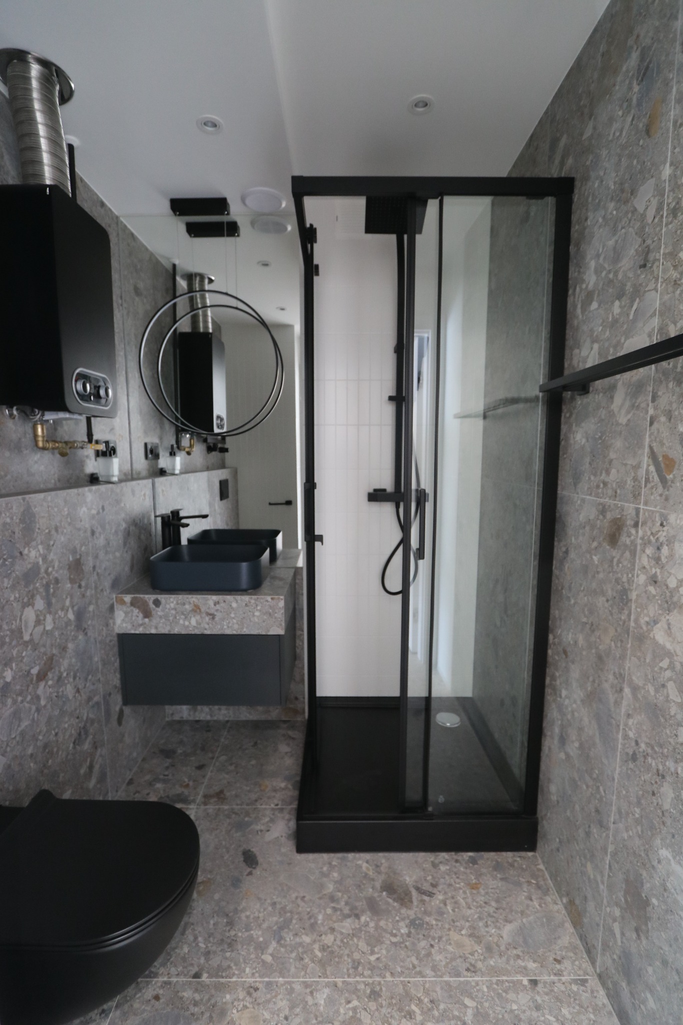 Nowoczesna łazienka z czarną kabiną prysznicową, umywalką nablatową i wiszącą toaletą, wykończona płytkami imitującymi lastryko, z widocznym grzejnikiem i okrągłym lustrem.