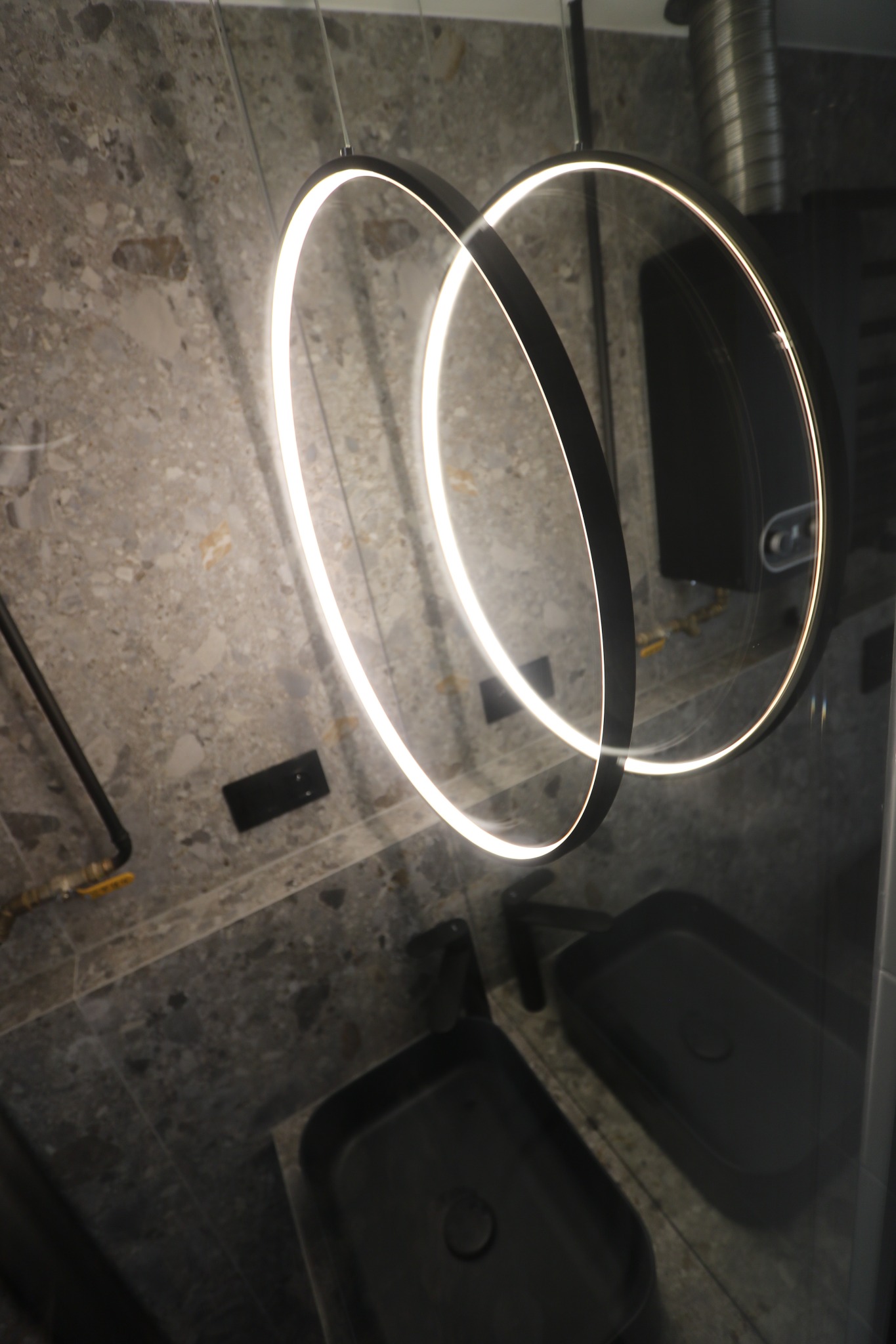 Nowoczesna łazienka z dwoma czarnymi umywalkami, podświetlanym okrągłym lustrem i marmurową ścianą.