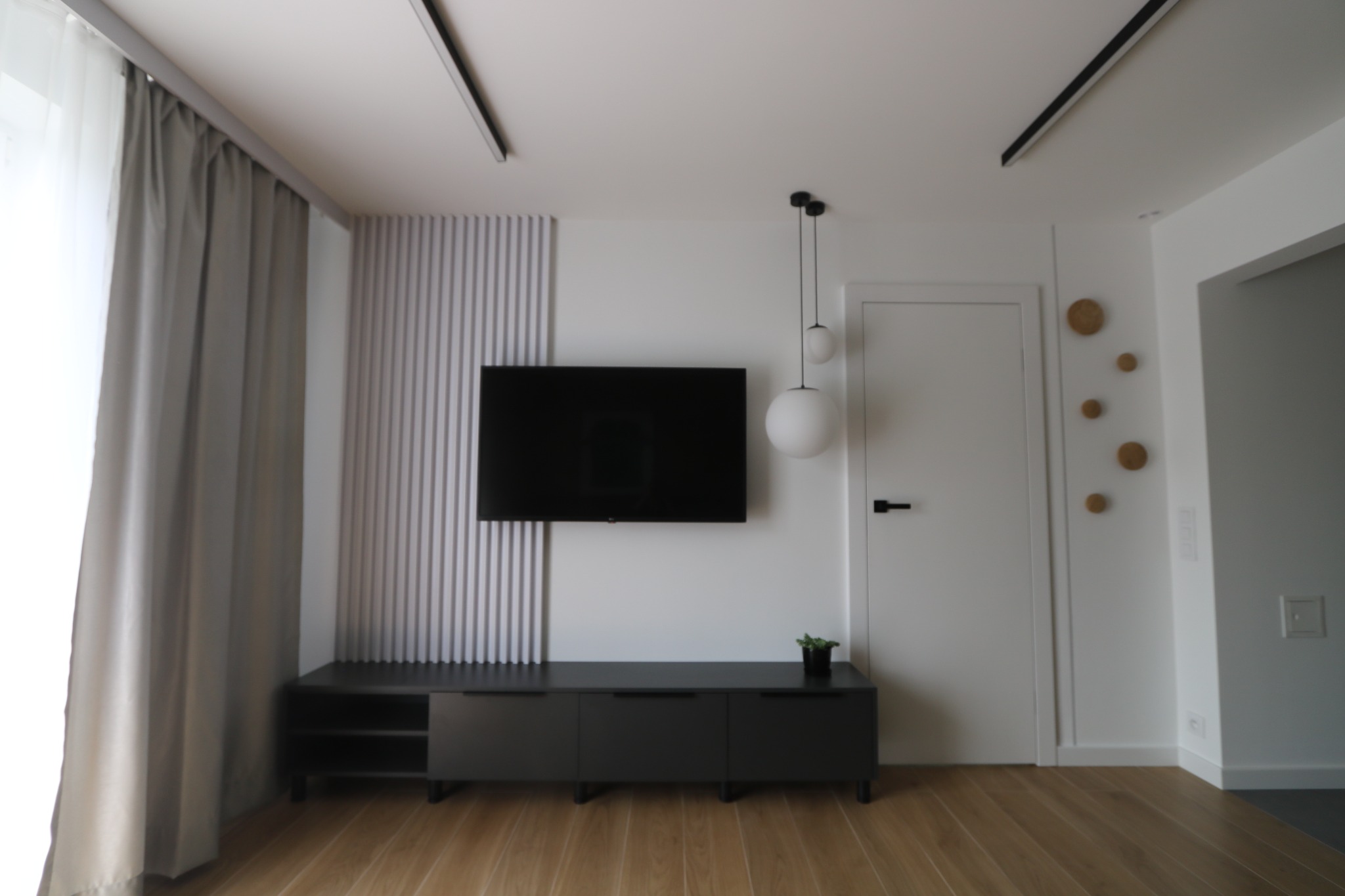 Minimalistyczny salon z zawieszonym telewizorem, czarną szafką RTV, panelami ściennymi z pionowych listew i drewnianą podłogą w odcieniach brązu