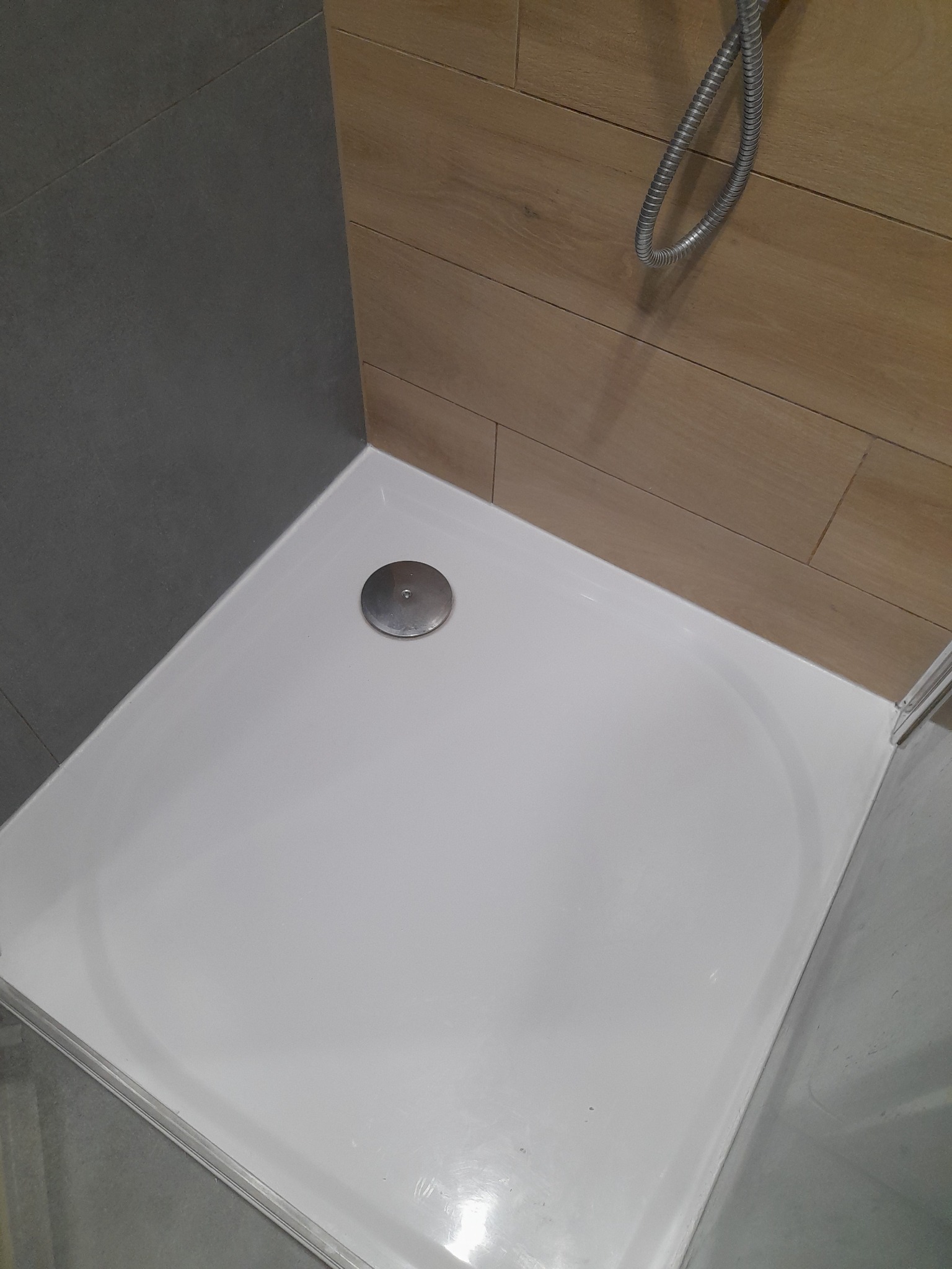 Biały brodzik z odpływem w narożnej kabinie prysznicowej, ściany wyłożone szarą płytką i drewnopodobną okładziną, widoczny wąż prysznicowy.