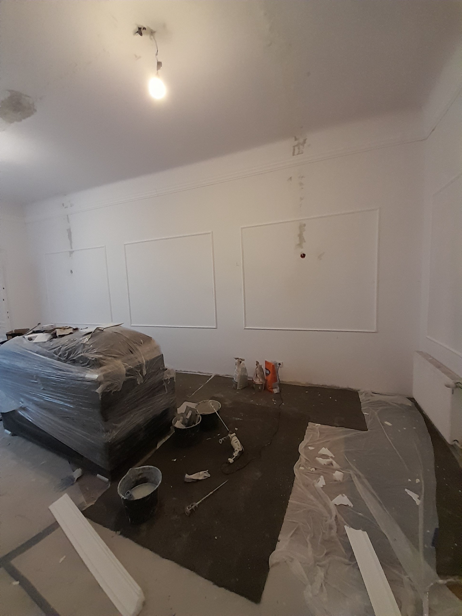 Pokój w trakcie prac wykończeniowych: ściany pomalowane na biało z dekoracyjnymi listwami, podłoga zabezpieczona folią, meble przykryte folią ochronną, widoczne narzędzia malarskie i wiadro z farbą.