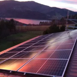 Panele słoneczne na czerwonym dachu w promieniach zachodzącego słońca, w tle górski krajobraz z jeziorem.