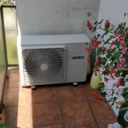 Zewnętrzna jednostka klimatyzacji marki HEIKO na balkonie z terakotą, otoczona kwitnącymi pelargoniami w donicach, widok z boku.