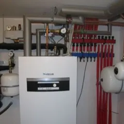 Biały piec grzewczy Buderus z rozbudowaną instalacją rur miedzianych i czarnych, manometry, zbiorniki wyrównawcze, rozdzielacz grzejnikowy z licznymi zaworami, zamontowany na jasnej ścianie...