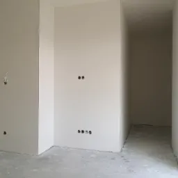 Wnętrze w trakcie remontu: ściany pokryte gładzią, widoczne puszki elektryczne, surowa podłoga, przejście do kolejnego pomieszczenia.