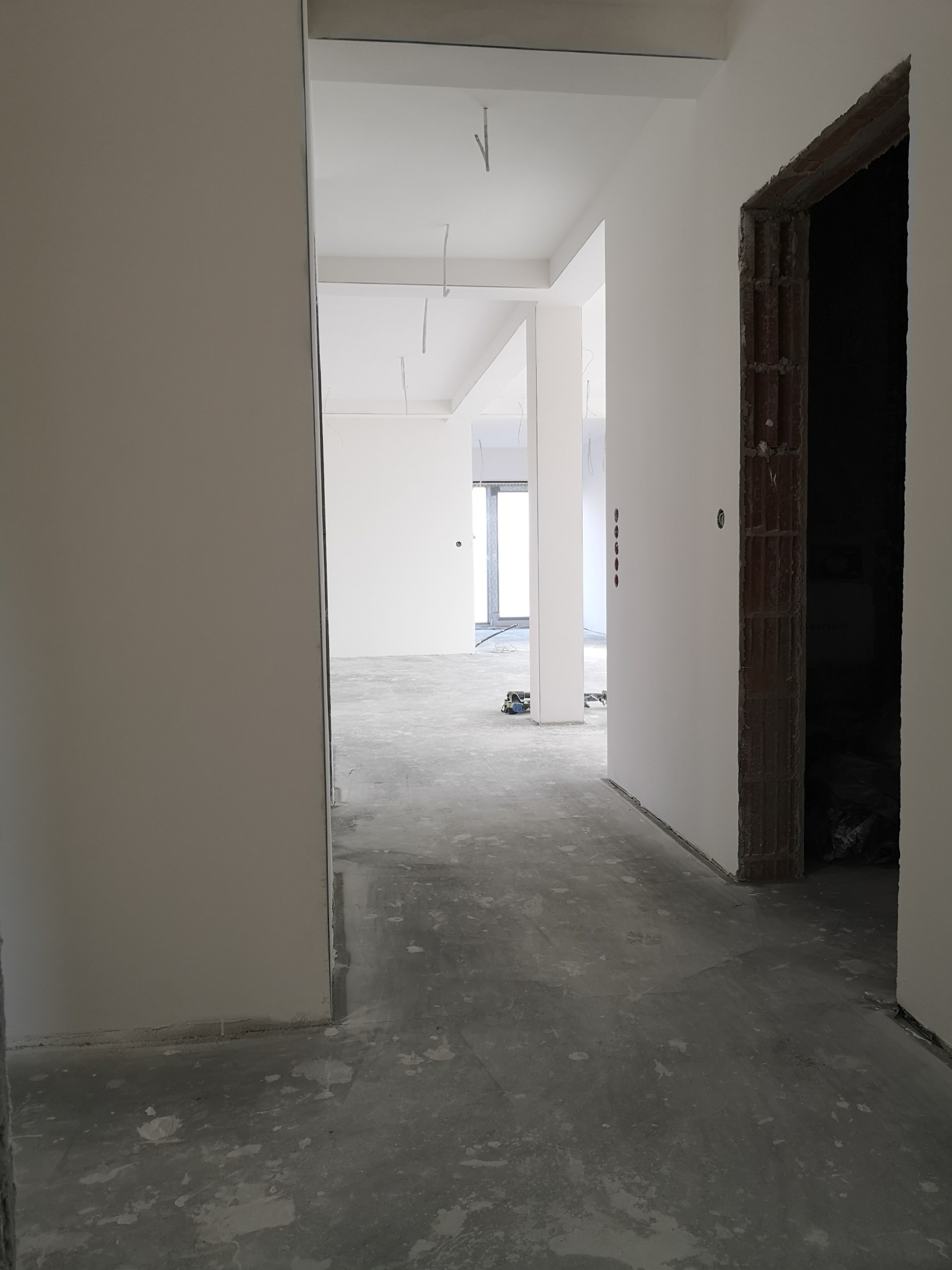 Wnętrze w trakcie remontu: surowe, białe ściany, betonowa podłoga z plamami farby, widoczne otwory po gniazdkach elektrycznych, niedokończone ościeżnice i elementy instalacji pod sufitem.