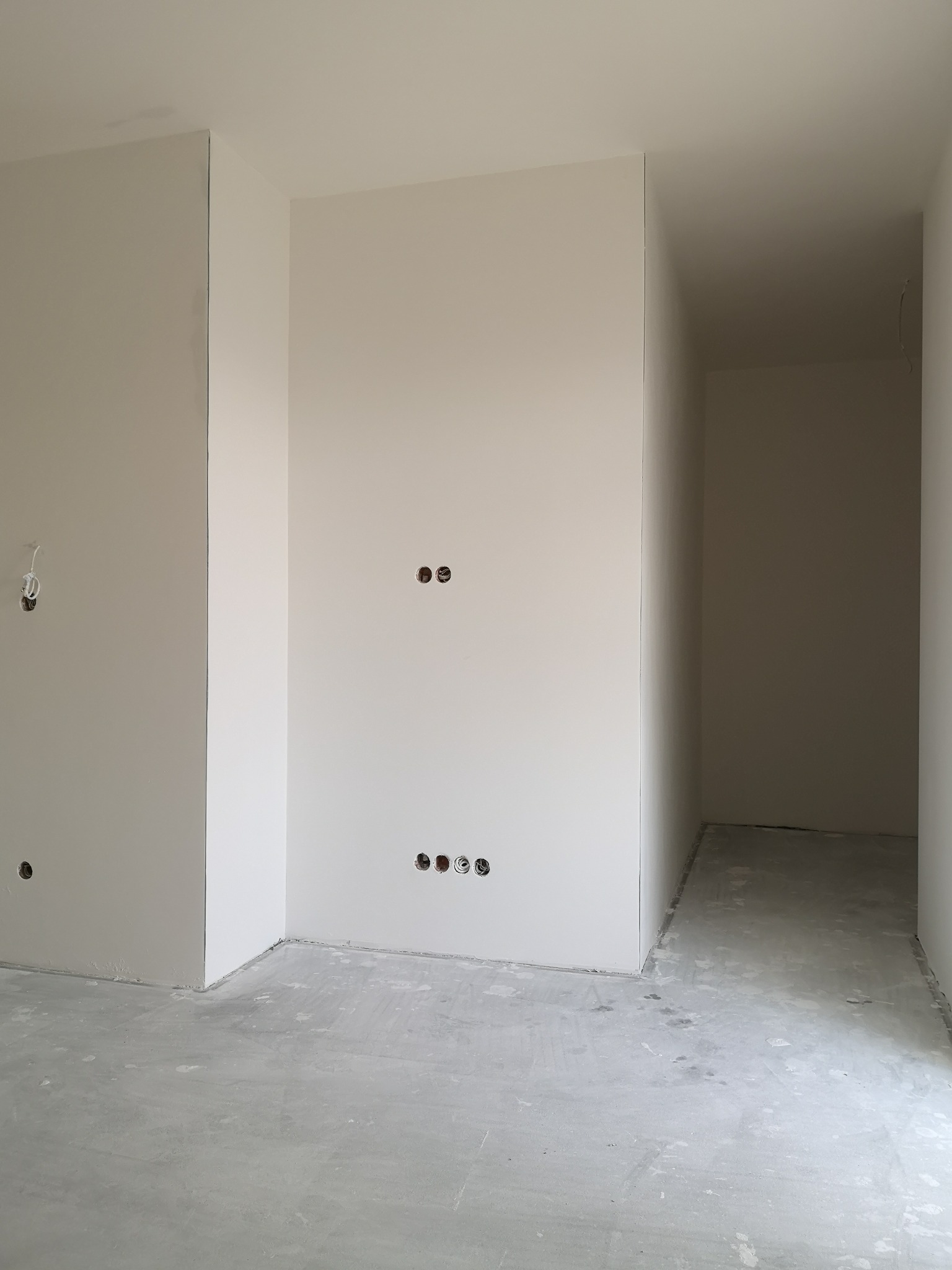 Wnętrze w trakcie remontu: ściany pokryte gładzią, widoczne puszki elektryczne, surowa podłoga, przejście do kolejnego pomieszczenia.