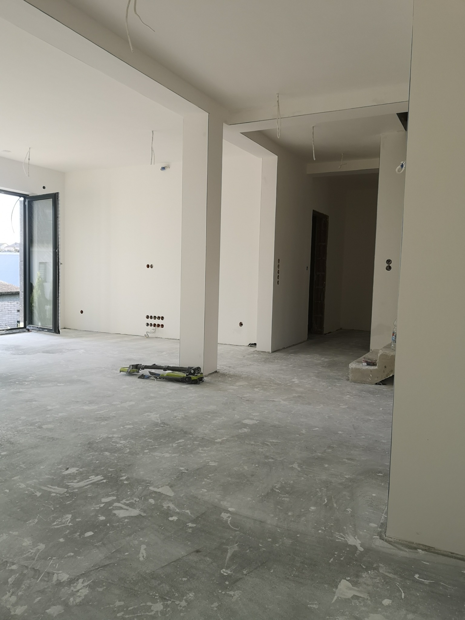 Wnętrze w trakcie prac wykończeniowych: surowy beton na podłodze z plamami farby, białe ściany z widocznymi puszkami elektrycznymi, narzędzie do szlifowania na podłodze, otwarte drzwi balkonowe w tle.