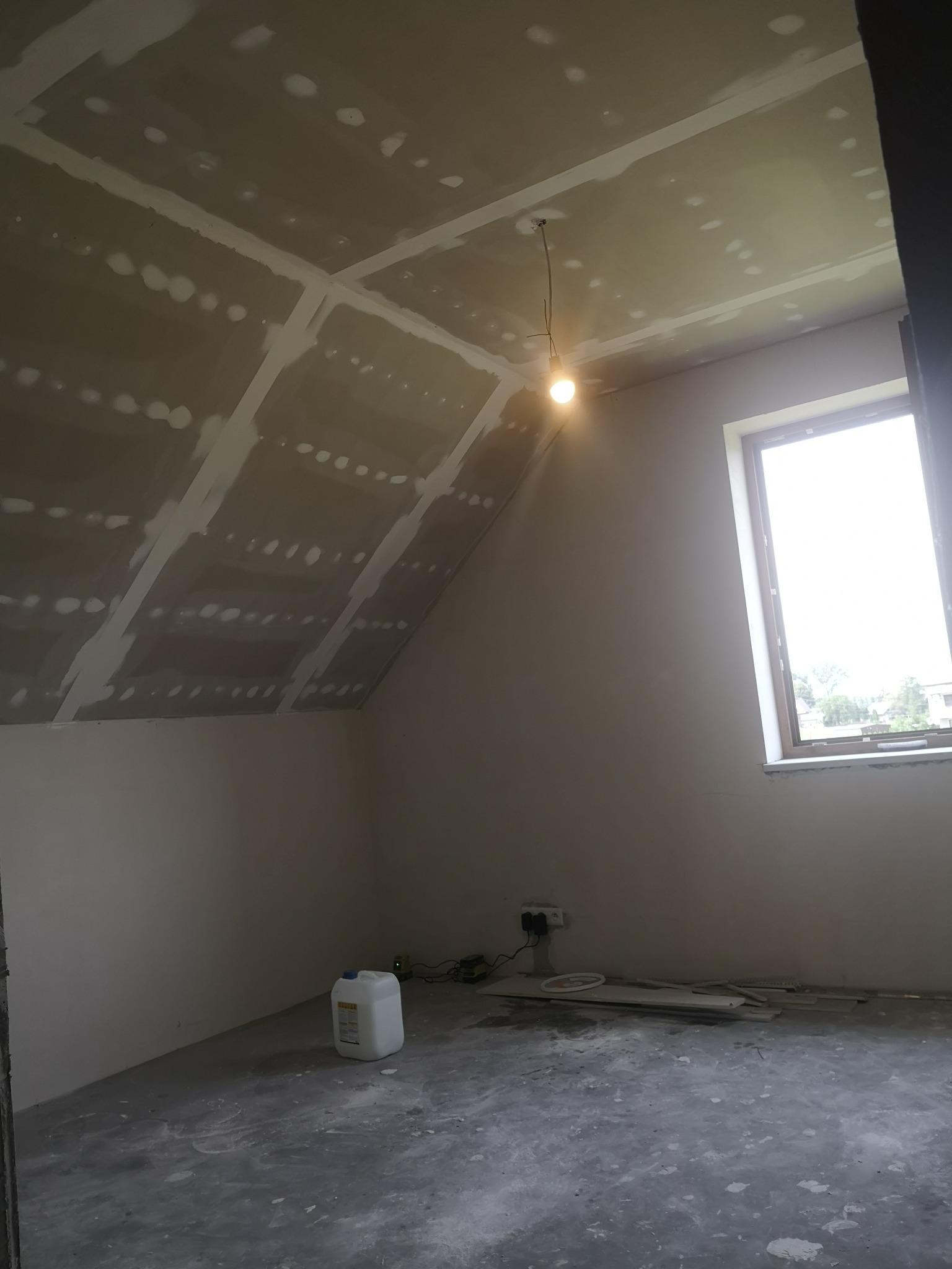 Pokój na poddaszu w trakcie remontu, z zamontowaną płytą kartonowo-gipsową na skosach sufitu, widoczne łączenia płyt i szpachlowanie, surowa żarówka zwisająca z sufitu, okno w tle i plastikowy...