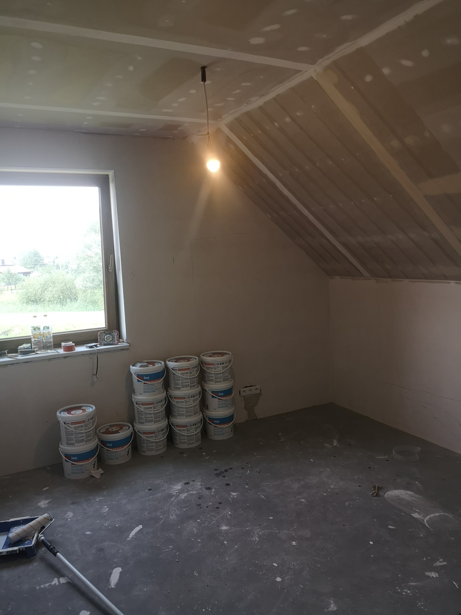 Poddasze w trakcie prac wykończeniowych: ściany przygotowane pod malowanie, widoczne szpachlowane płyty karton-gips na skosach, wiaderka z farbą Knauf Grand Finish oraz wałek malarski na podłodze...