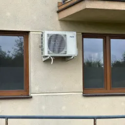 Biały klimatyzator AUX zamontowany na elewacji budynku, widoczny między dwoma oknami. Ujęcie z dołu, z fragmentem balustrady na pierwszym planie.