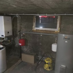 W.H GROUP Sp. z o.o. - Piwnica z zamontowanym kotłem gazowym Kaisai, zasobnikiem Ferroli, instalacją hydrauliczną i oknem. Widoczne rury i akcesoria. Ściany z cegły i betonu.