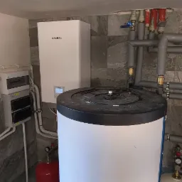 Instalacja Kaisai z zbiornikiem, inwerterem PV FoxEss i rurami w szarym oplocie na tle ściany z szarej mozaiki. Elementy instalacji grzewczej w kotłowni.