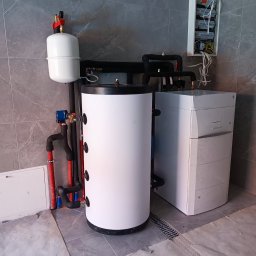 W.H GROUP Sp. z o.o. - Biały zasobnik ciepłej wody użytkowej z podłączonymi rurami, obok piec gazowy w folii ochronnej, na tle szarych płytek. Widoczna skrzynka elektryczna.
