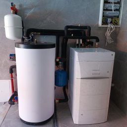 W.H GROUP Sp. z o.o. - Nowoczesna kotłownia z białym zasobnikiem ciepłej wody i piecem gazowym De Dietrich, czarne rury izolacyjne, szara płytka na ścianie i podłodze, skrzynka elektryczna.