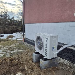 W.H GROUP Sp. z o.o. - Biała pompa ciepła Kaisai zamontowana na betonowych blokach przy ścianie budynku z widoczną izolacją. W tle resztki śniegu i drzewa.