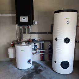 W.H GROUP Sp. z o.o. - Zestaw urządzeń grzewczych: czarny zasobnik, biały zbiornik z fakturą plastra miodu, biały zbiornik z osprzętem, połączone rurami w szarej kotłowni.