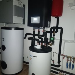 W.H GROUP Sp. z o.o. - Zestaw urządzeń grzewczych: czarny piec z wyświetlaczem, czerwony zbiornik wyrównawczy, białe zasobniki wody. Rury, zawory i manometr. Widok z niskiego kąta.