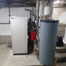 W.H GROUP Sp. z o.o. - Nowoczesna instalacja grzewcza w piwnicy: biały piec, szary zasobnik wody, czerwony zbiornik wyrównawczy, rury i zawory. Widoczne schody i surowe ściany.