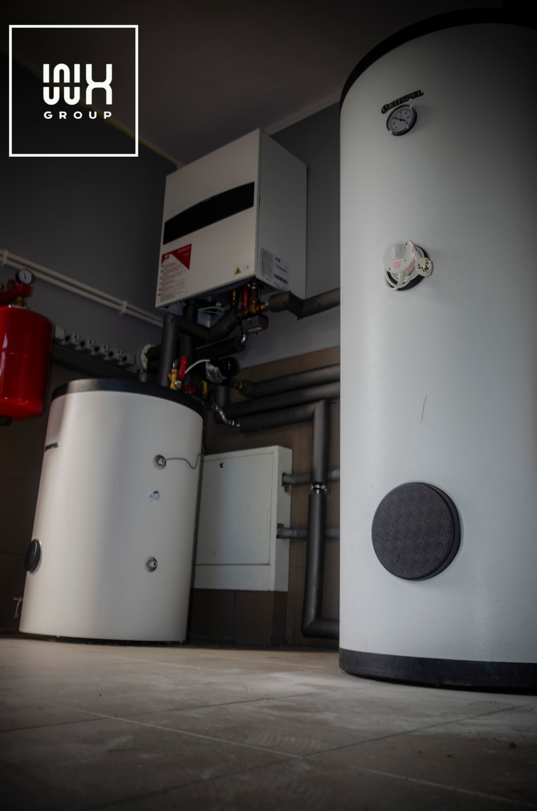 Biały, wiszący piec gazowy i dwa białe, cylindryczne zbiorniki akumulacyjne z rurami w szarym pomieszczeniu technicznym, widok z dołu.