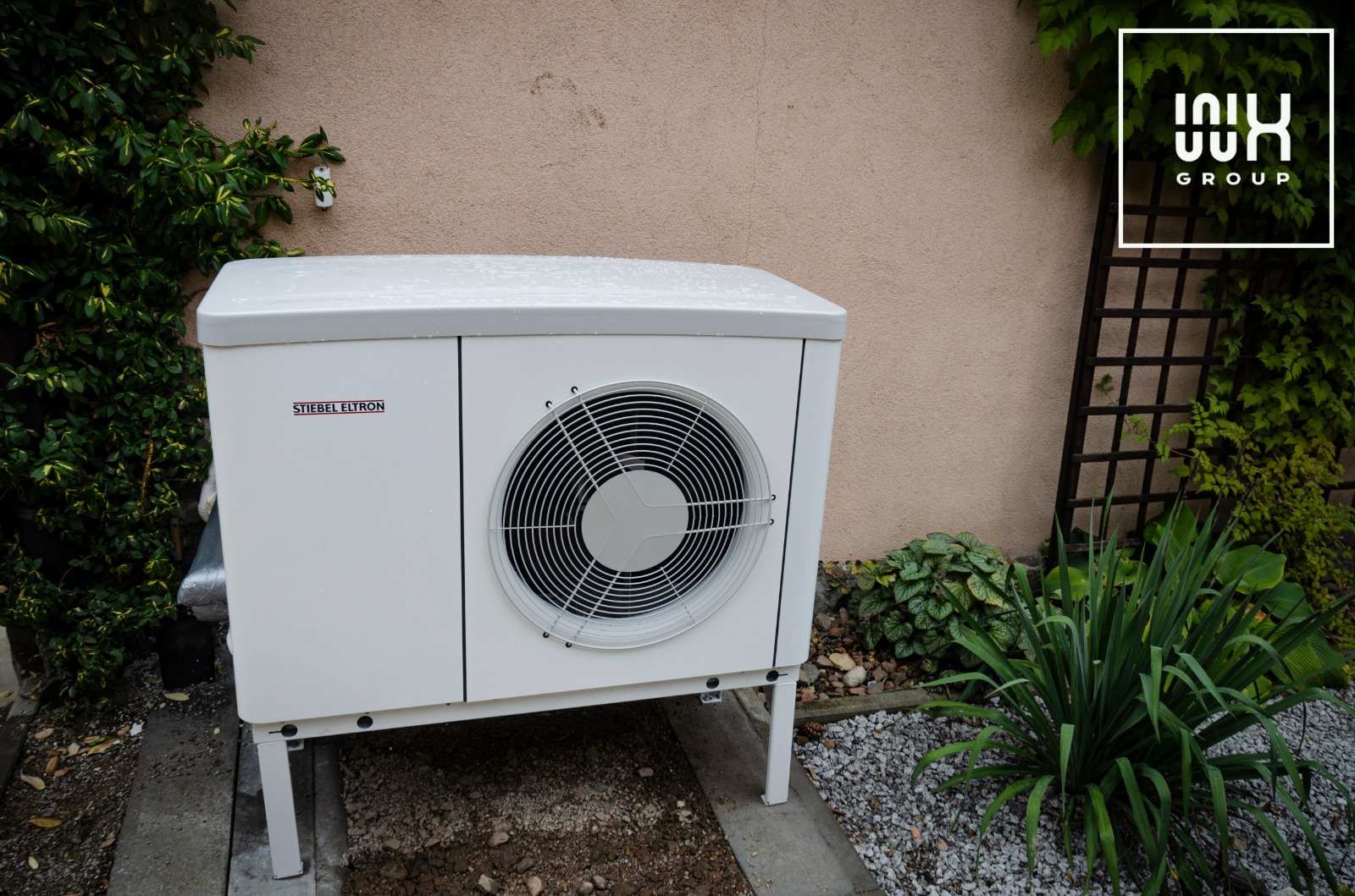 Biała pompa ciepła Stiebel Eltron na tle beżowej ściany i zieleni. Widoczny fragment logo firmy. Urządzenie stoi na żwirowym podłożu.